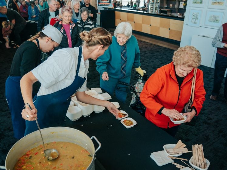 Image of A chef serves soup into bowls for a queue of elderly people at a community event. Attendees wait with trays and cutlery, while others chat in the background. The setting appears bright and welcoming.