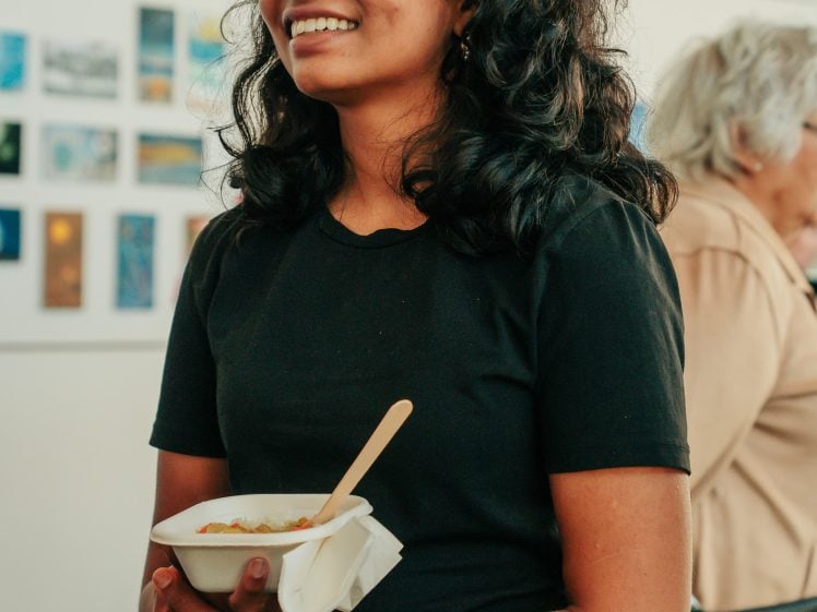 Image of A woman with curly dark hair, wearing a black shirt, smiles while holding a bowl of food and a wooden spoon. There are small artworks displayed on the wall behind her, and another person is blurred in the background.