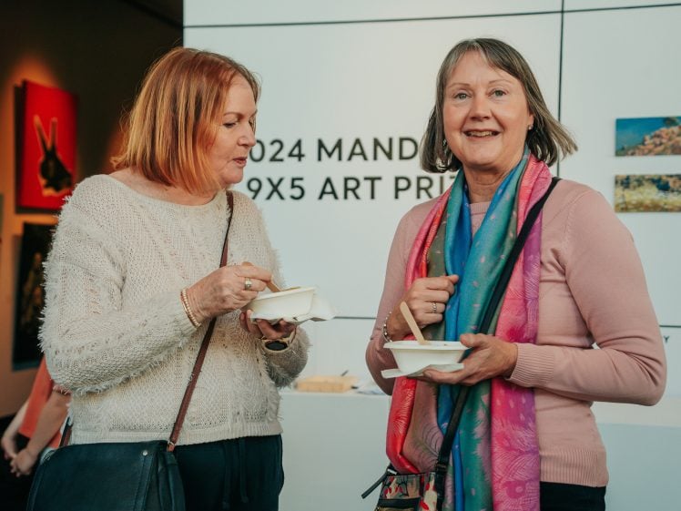 Image of Two women standing indoors, holding bowls of food and smiling. They are dressed in casual jumpers and scarves. Artworks are displayed on the wall behind them, along with a sign reading 2024 MAND... 9X5 ART PRI....