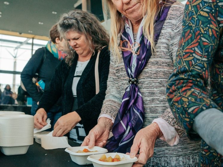 Image of A woman in a purple scarf serves food from a container at a table, with other people standing beside her, also serving food. The setting appears to be indoors, possibly at a community event.