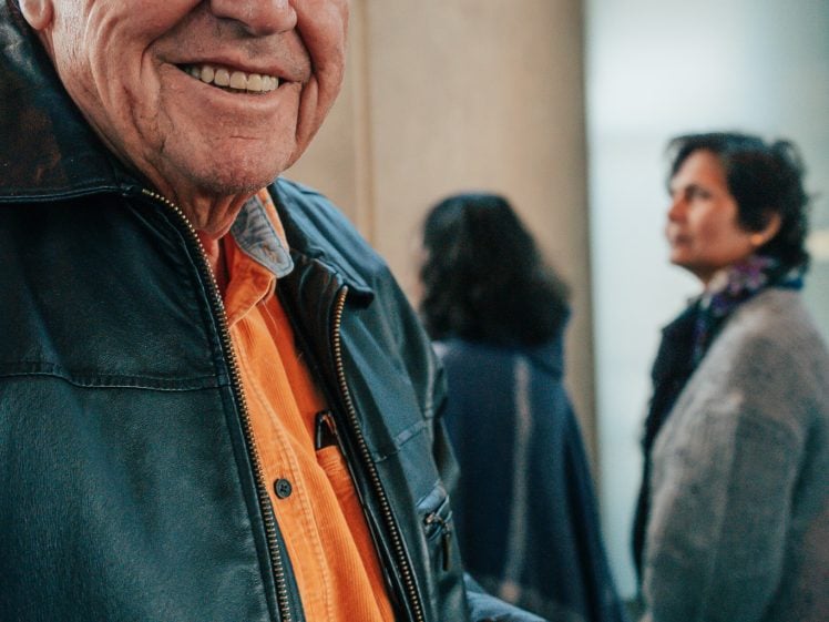 Image of An older man in a black hat and leather jacket smiles whilst holding a bowl of soup with a spoon. Two people stand blurred in the background. The setting appears to be indoors.