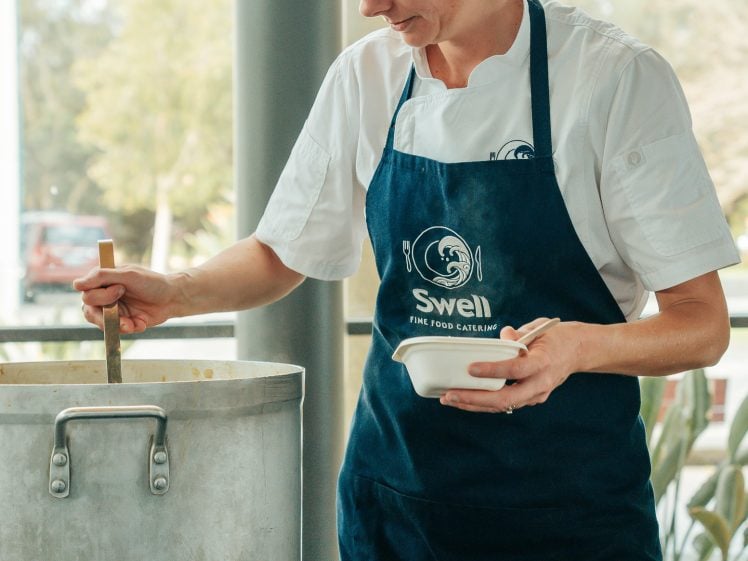 Image of A chef wearing a white shirt and a navy blue apron stirs a large pot with a wooden spoon while holding a small bowl, standing in a bright kitchen with windows and plants in the background.