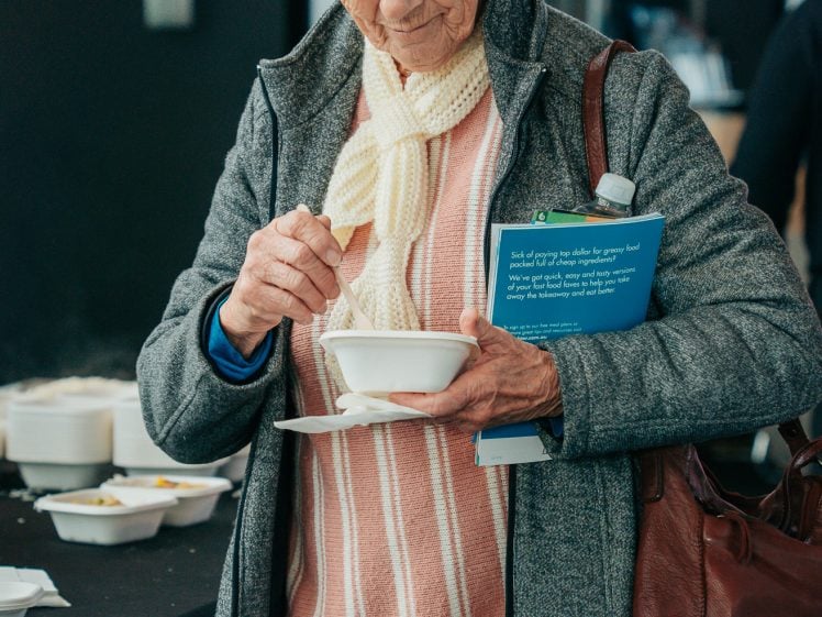 Image of An older woman wearing glasses, a white scarf, and a grey coat holds a book and stirs a bowl of soup while standing indoors. She carries a brown bag and appears focused on her food.