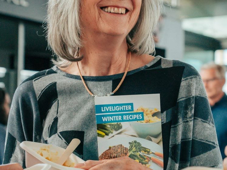 Image of An older woman with grey hair and glasses smiles whilst holding a LiveLighter Winter Recipes booklet. People around her hold food containers. The setting appears to be indoors, possibly at an event or gathering.