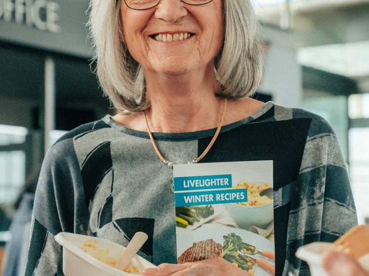 Image of An older woman with grey hair and glasses smiles whilst holding a LiveLighter Winter Recipes booklet and some food in a bright indoor setting.