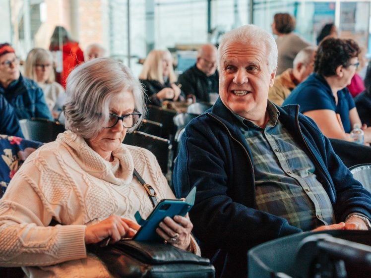 Image of An older woman in a cream jumper uses a tablet while sitting next to a smiling older man in a checked shirt and jacket. They are seated among a group of people in what appears to be a waiting area.