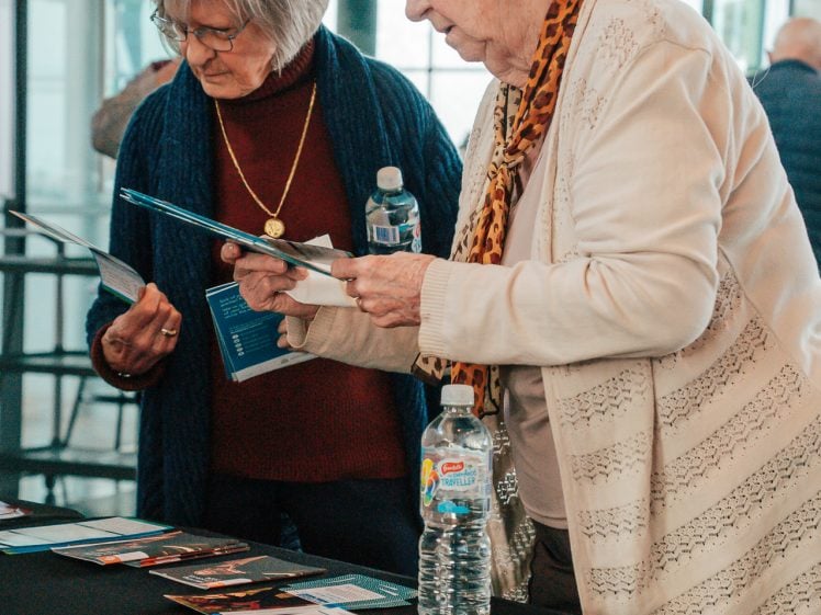 Image of Two elderly women stand at a table covered with leaflets and brochures, looking through the materials. Both hold water bottles; one wears a blue jumper and the other a cream cardigan with a leopard scarf.