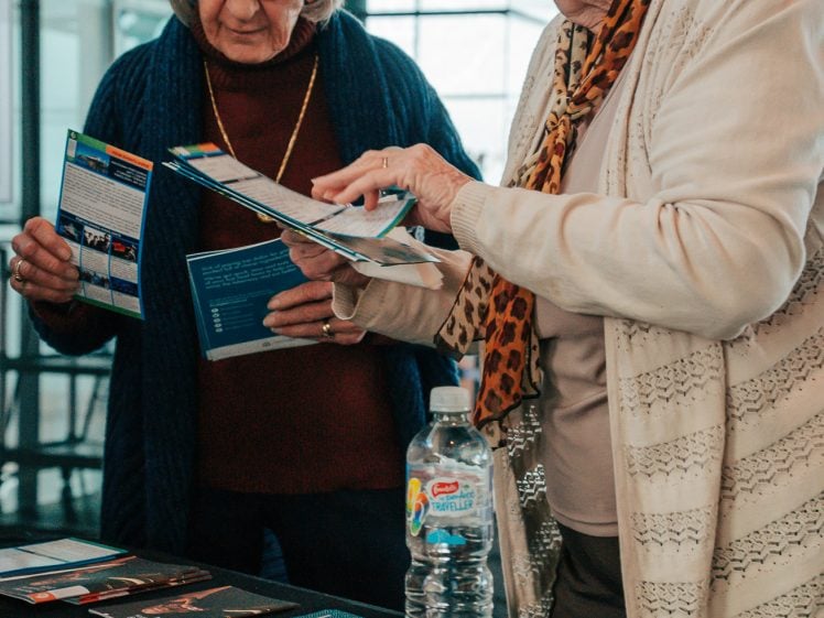 Image of Two elderly women stand at a table covered with pamphlets and brochures, reading and discussing the materials. A water bottle is also on the table. They appear engaged and focused in a bright indoor setting.