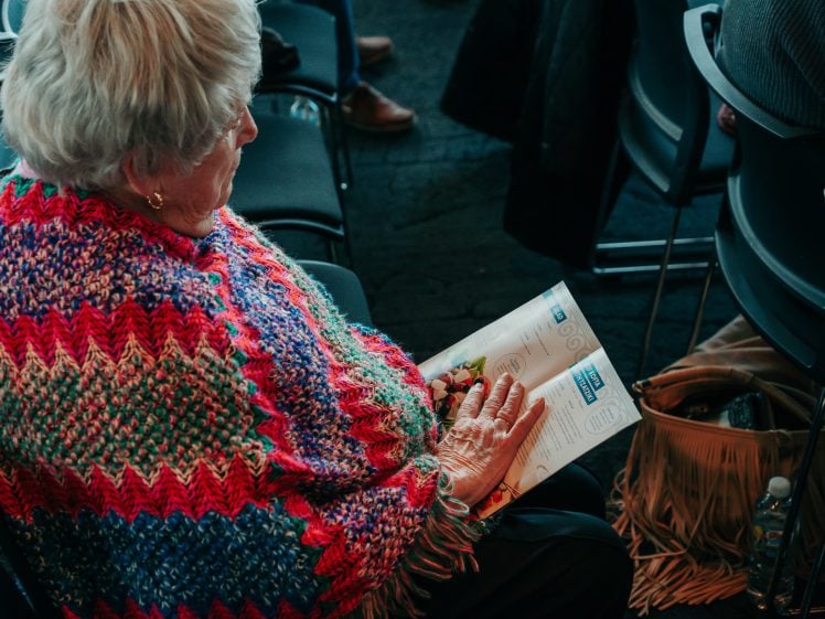 Image of An elderly person with white hair, wearing a colourful crocheted shawl, sits among rows of chairs and reads a magazine or programme. A tan fringed bag and a water bottle are on the floor beside their seat.
