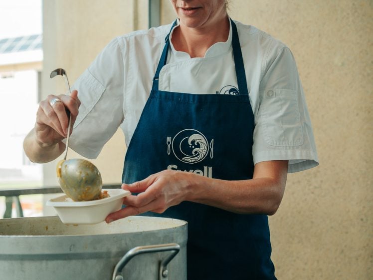 Image of A chef wearing a white shirt and blue apron ladles soup from a large pot into a bowl, standing indoors near a window.
