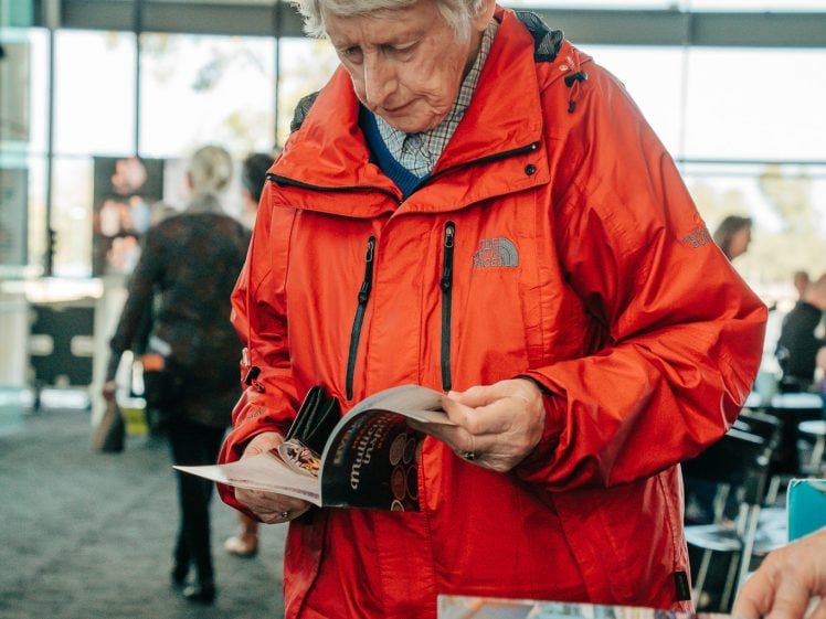 Image of An older adult with short grey hair, wearing a bright red jacket, stands indoors and looks closely at a booklet. Other people and tables with magazines are visible in the background.