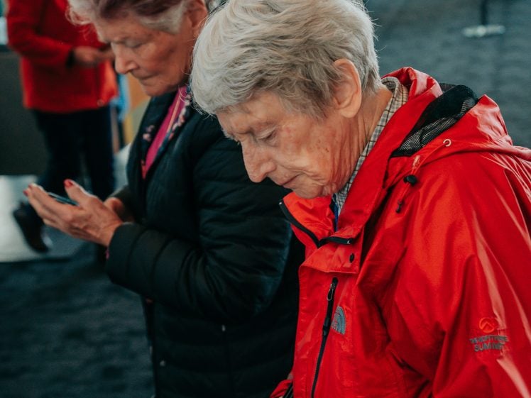 Image of Two elderly women stand indoors, one in a red jacket reading a leaflet, the other in a black jacket looking down at her mobile, with blurred people and displays in the background.