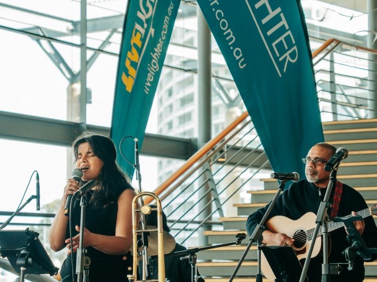 Image of A woman sings into a microphone beside a trombone, while a man plays guitar on stage. Behind them are LiveLighter banners and stairs; musical equipment is visible in the modern indoor setting.