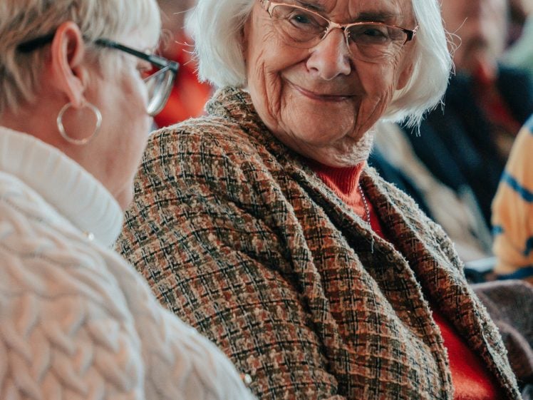 Image of Two older women sit next to each other in conversation. One, with white hair, glasses, and a checked jacket, smiles warmly at the other, who has short blonde hair and wears a white jumper. There are people blurred in the background.