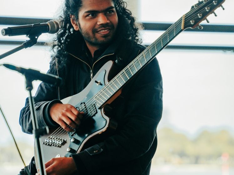 Image of A person with long curly hair sits on a stool, holding an electric guitar and smiling, with a microphone positioned nearby. The background shows an outdoor scene with trees and water.