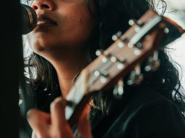 Image of A person with long, wavy dark hair plays an acoustic guitar and sings into a microphone, looking intently at the camera with a focused expression.