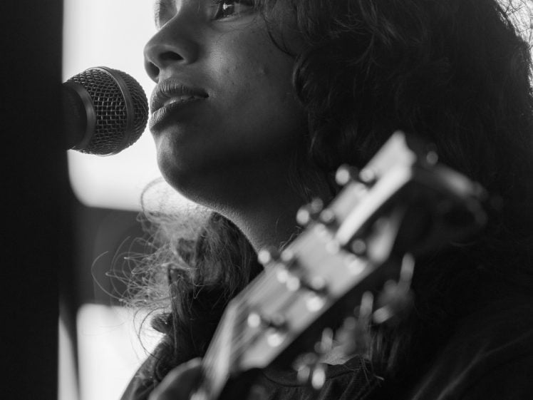 Image of Black and white photo of a person with long curly hair singing into a microphone while playing an acoustic guitar, shown in close-up from the side.