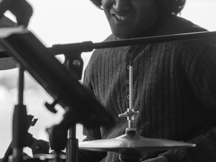 Image of A person with medium-length hair plays the drums with intensity, surrounded by microphones and music equipment. The image is in black and white, capturing a moment of musical focus and emotion.