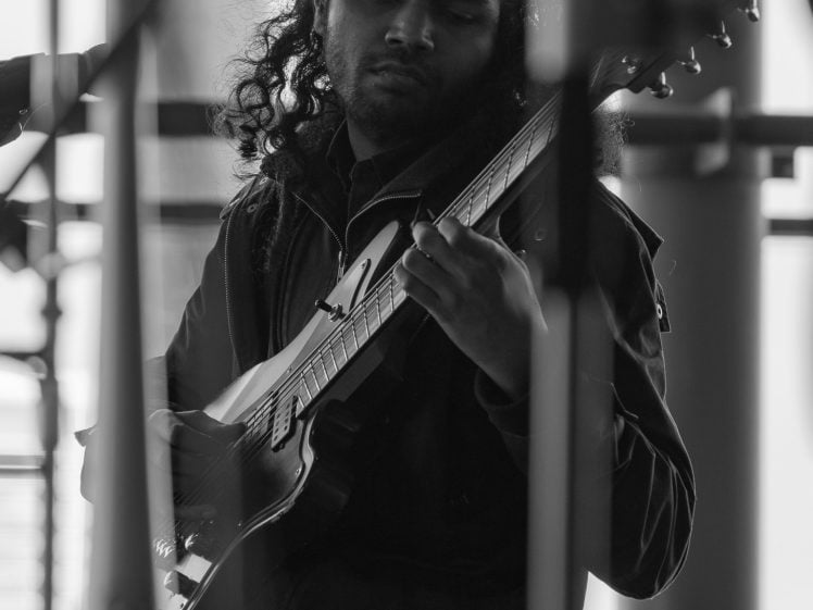 Image of A musician with long curly hair plays an electric guitar while seated. The image is in black and white, with microphones and a blurred trombone visible in the foreground.