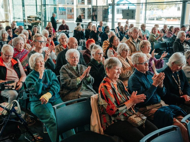 Image of A large group of older adults sit closely together in chairs, some clapping and smiling, at an indoor event with large windows and posters in the background.