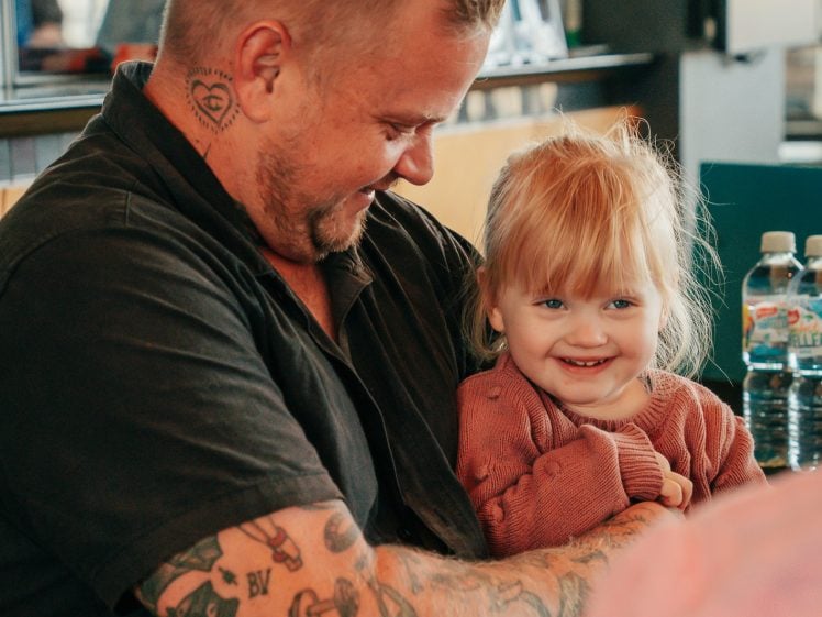 Image of A smiling man with tattoos and short hair holds a happy, blonde toddler in his lap. They are indoors, sitting closely together, and both look joyful. Bottled water and other items are visible in the background.