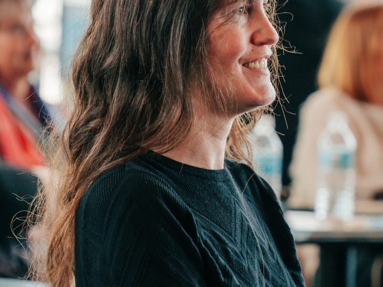 Image of A woman with long, wavy brown hair and a black jumper smiles whilst sitting indoors, surrounded by blurred people and water bottles in the background.