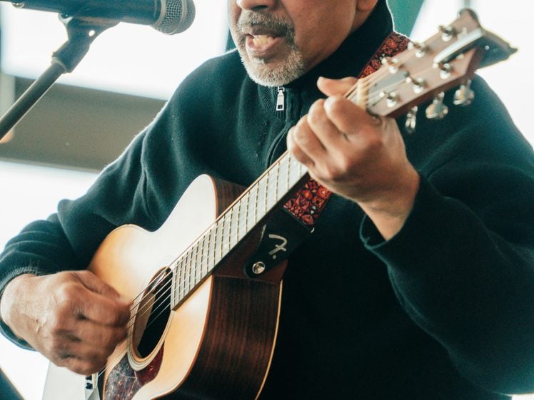 Image of A man wearing glasses and a black jumper plays an acoustic guitar and sings into a microphone indoors, with sunlight streaming through windows behind him.