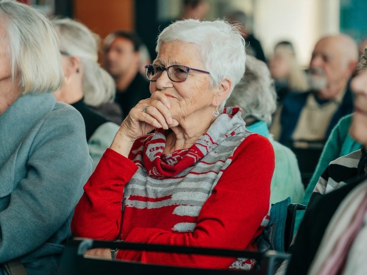 Image of An elderly woman with short white hair, glasses, and a red jumper with a grey scarf sits among a group of older adults, looking thoughtful and attentive during an event or gathering.