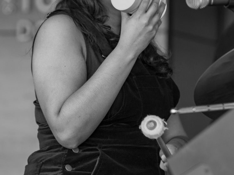 Image of A woman sits at a drum kit, holding a mug close to her mouth as if about to drink, with a microphone positioned nearby. The image is in black and white.