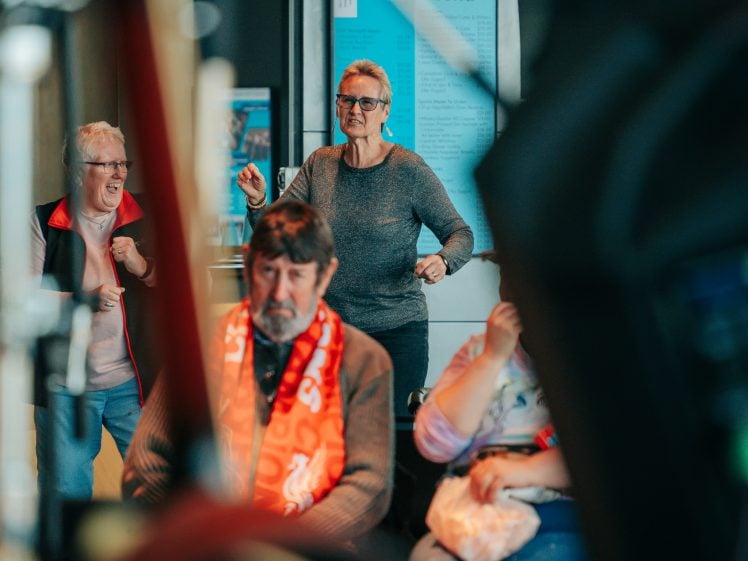 Image of Two older women stand smiling and dancing in the background, whilst a man in a red scarf and another seated person sit in the foreground, partially obscured. A blue drinks menu is visible on the wall behind them.
