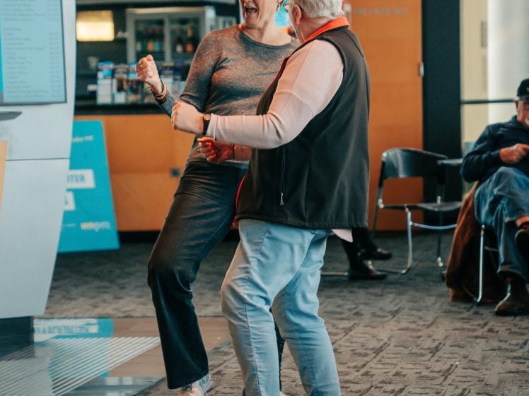 Image of Two older adults are smiling and dancing together indoors, facing each other with joy. A man is seated in the background, appearing relaxed. The scene is casual and lively, with natural light coming through windows.