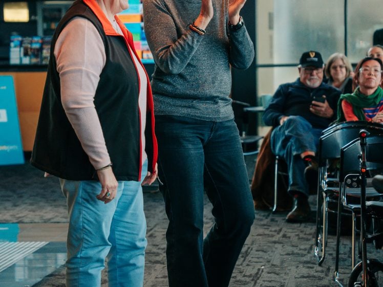 Image of Two smiling older women stand indoors, one clapping whilst the other faces the audience. Several people sit and watch from the background in a modern, well-lit room.