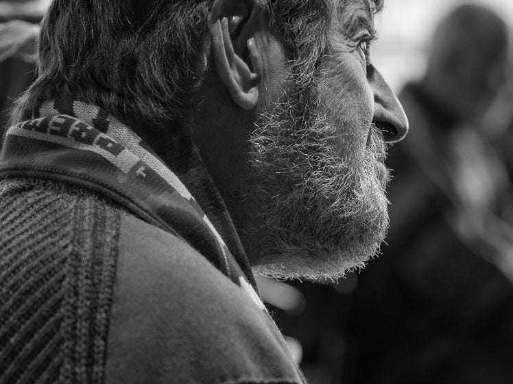 Image of A black and white photo of an older man with a beard and moustache, gazing thoughtfully to the right. He wears a textured jumper and scarf, with soft light illuminating his profile. The background is blurred.