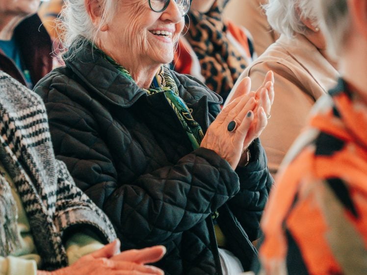 Image of An older woman with glasses and white hair in a bun smiles and claps whilst sitting among a group of people at an indoor event.