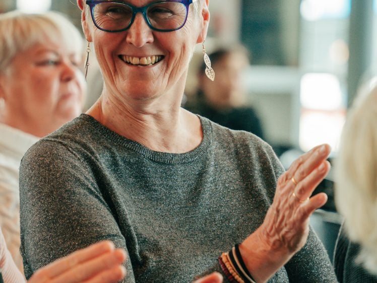 Image of An older woman with short grey hair and glasses smiles while clapping during an indoor event. She wears a grey top, earrings, and bracelets. Other people are seated and clapping in the background.