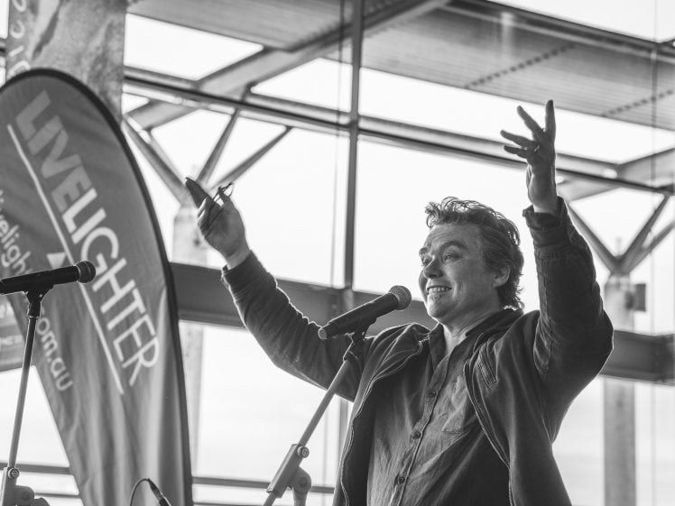 Image of A man stands in front of microphones with arms raised, appearing to speak enthusiastically. A LiveLighter banner is visible beside him. The setting is indoors with large windows in the background. The image is in black and white.