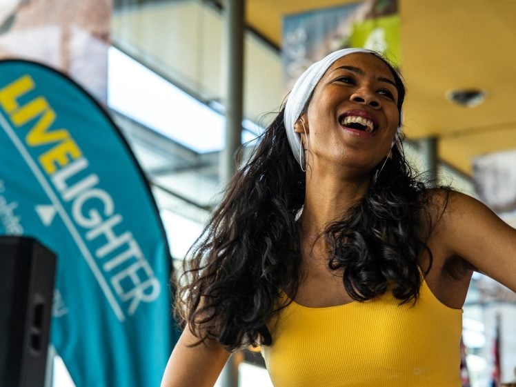 Image of A woman in a yellow top and white headband smiles brightly whilst dancing indoors. A teal LIVE LIGHTER banner is visible in the background.