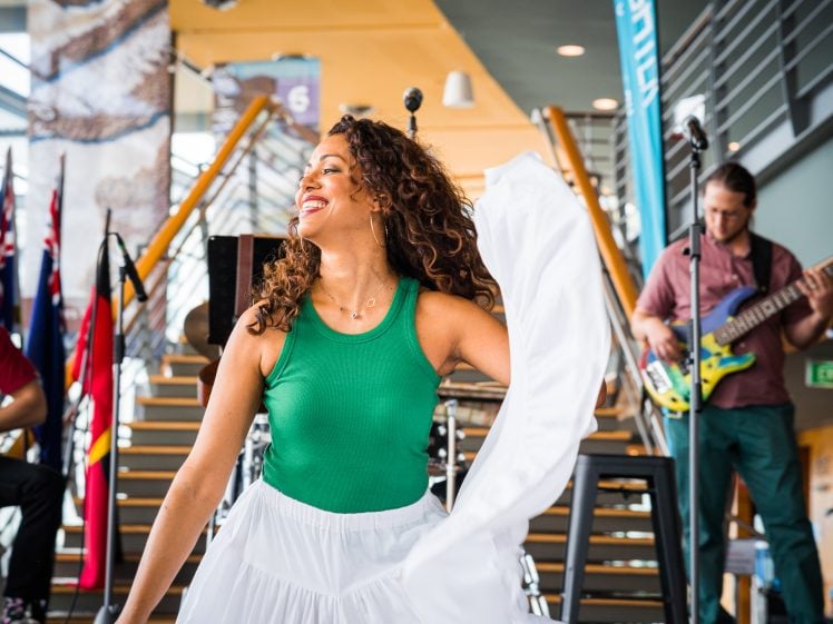 Image of A woman in a green top and white skirt dances joyfully, smiling with eyes closed, holding her skirt. Musicians play instruments beside her in a brightly lit indoor space with stairs and international flags in the background.