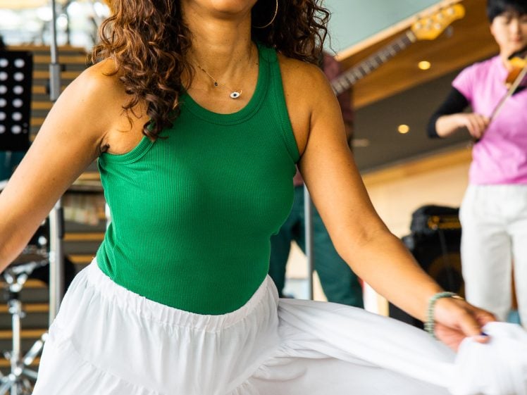 Image of A woman in a green sleeveless top and flowing white skirt dances joyfully indoors, smiling broadly. A musician in a pink shirt and white trousers plays guitar in the background.