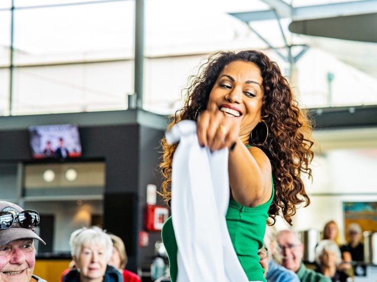 Image of A woman with curly hair, wearing a green top, smiles and dances whilst holding a white cloth in a bright room with seated people watching her.