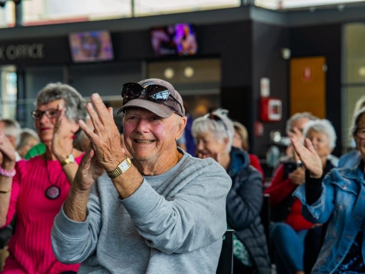 Image of A group of smiling older adults sit together indoors, enthusiastically clapping. The man in front wears a hat, sunglasses, and a grey jumper. The atmosphere appears joyful and lively.