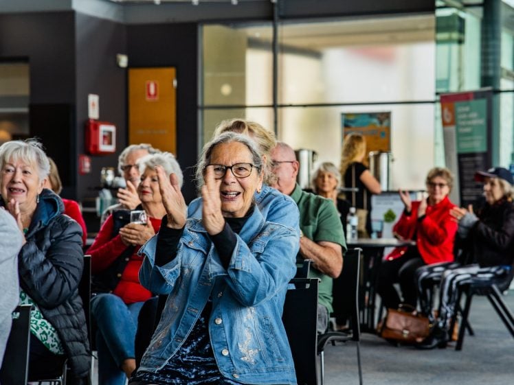 Image of A group of older adults sit indoors, smiling and clapping. The focus is on a woman in a denim jacket in the foreground, whilst others in colourful outfits are visible in the background, also applauding.