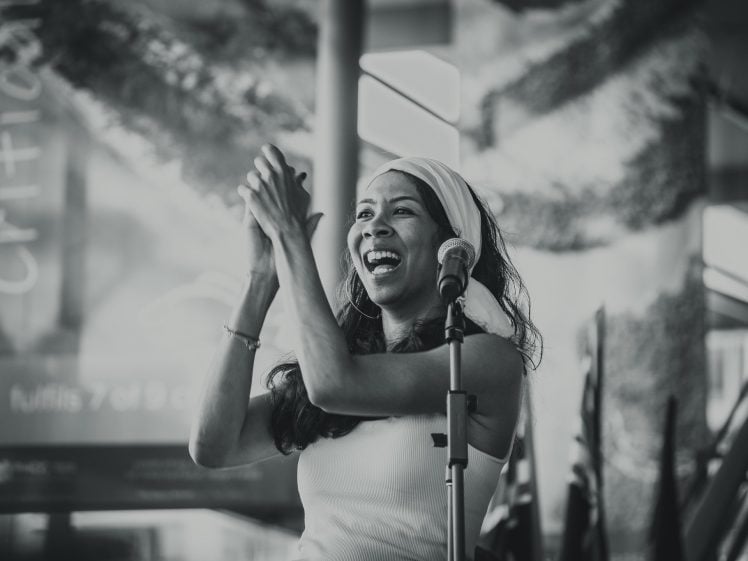 Image of A woman wearing a hairband smiles and claps while standing at a microphone, appearing to speak or perform in front of an audience. The photo is in black and white.