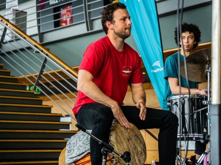 Image of A man in a red shirt plays a hand drum while seated, and another person in a blue shirt plays a drum kit behind him. They are indoors near a staircase, performing together.