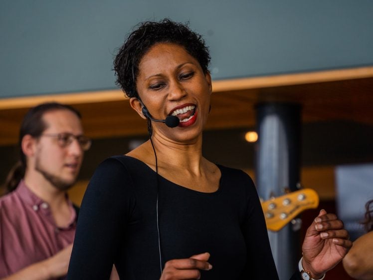 Image of A woman wearing a black top and headset microphone is singing and smiling with eyes closed. A man with glasses and a tied-back ponytail stands blurred in the background, and part of a guitar is visible.