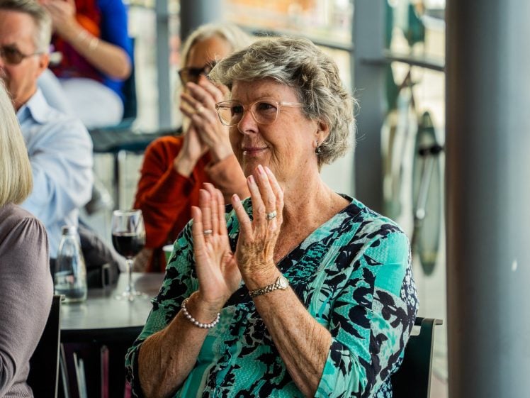 Image of An older woman with grey hair and glasses sits and claps, smiling warmly. She wears a blue patterned top and is surrounded by other people, some applauding, in a bright indoor setting. A glass of wine is on the table nearby.