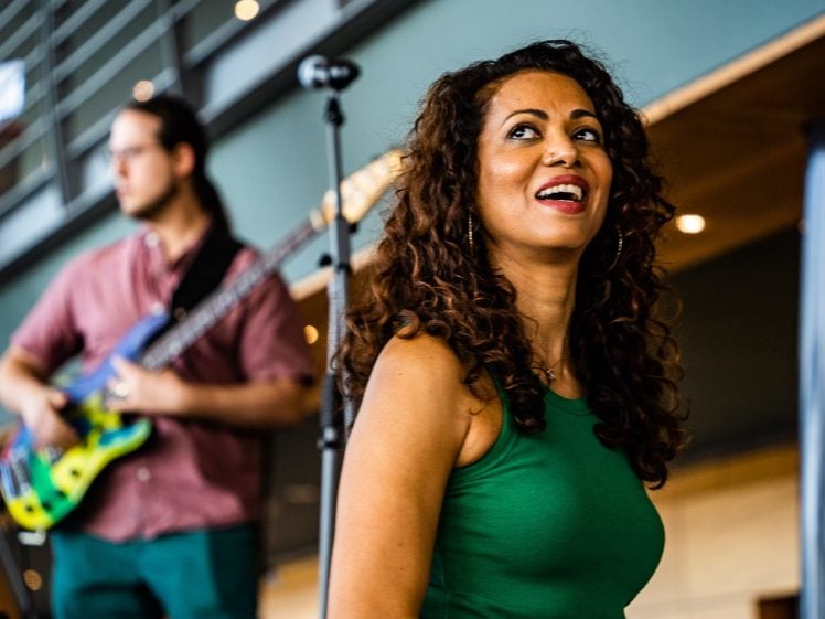 Image of A woman in a green sleeveless top smiles whilst standing near a microphone. In the background, a man plays an electric guitar. The setting appears to be indoors with modern décor and lighting.