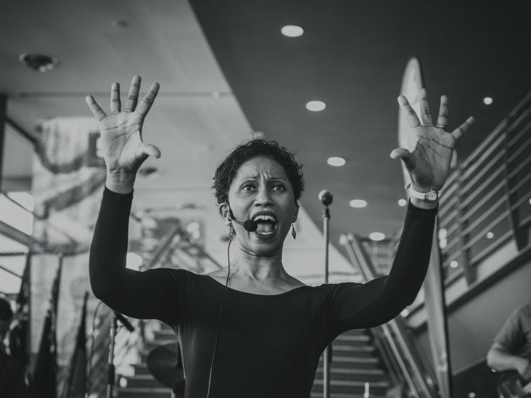 Image of A woman wearing a headset microphone stands indoors with her arms raised and fingers spread wide, appearing to sing or speak passionately. People and modern architecture are visible in the background.