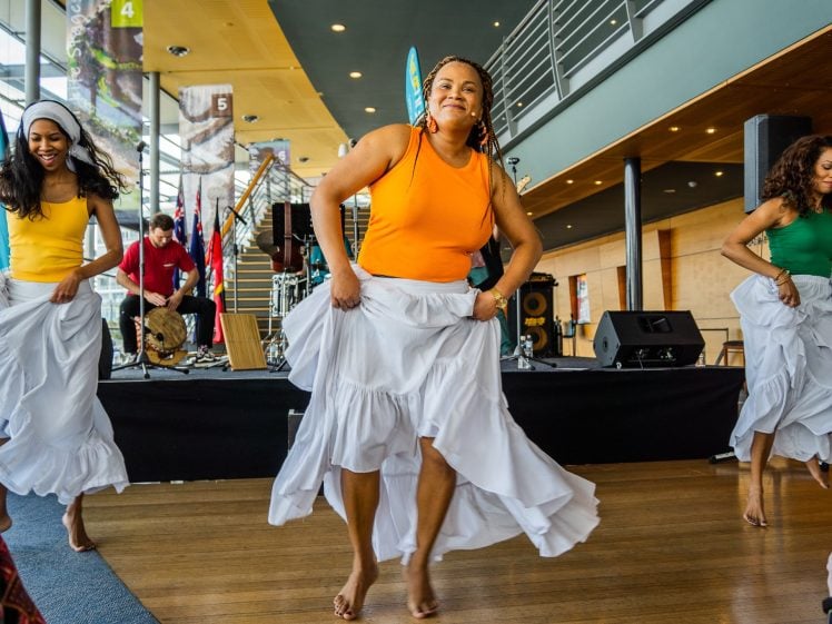 Image of Three women in colourful tops and white skirts dance barefoot on a wooden floor, smiling, with musicians and flags in the background in a bright indoor space.