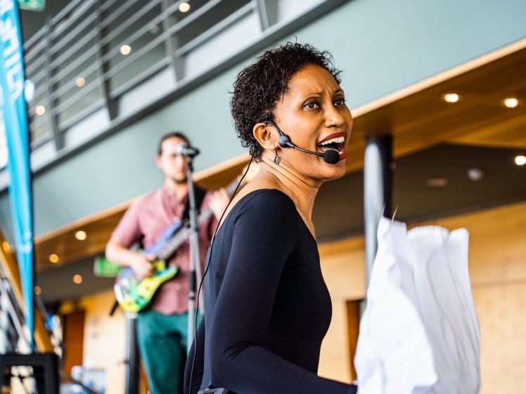 Image of A woman wearing a headset microphone smiles whilst performing indoors, holding white papers. In the background, a musician plays an electric guitar on stage. Modern railings and wood panelling are visible.